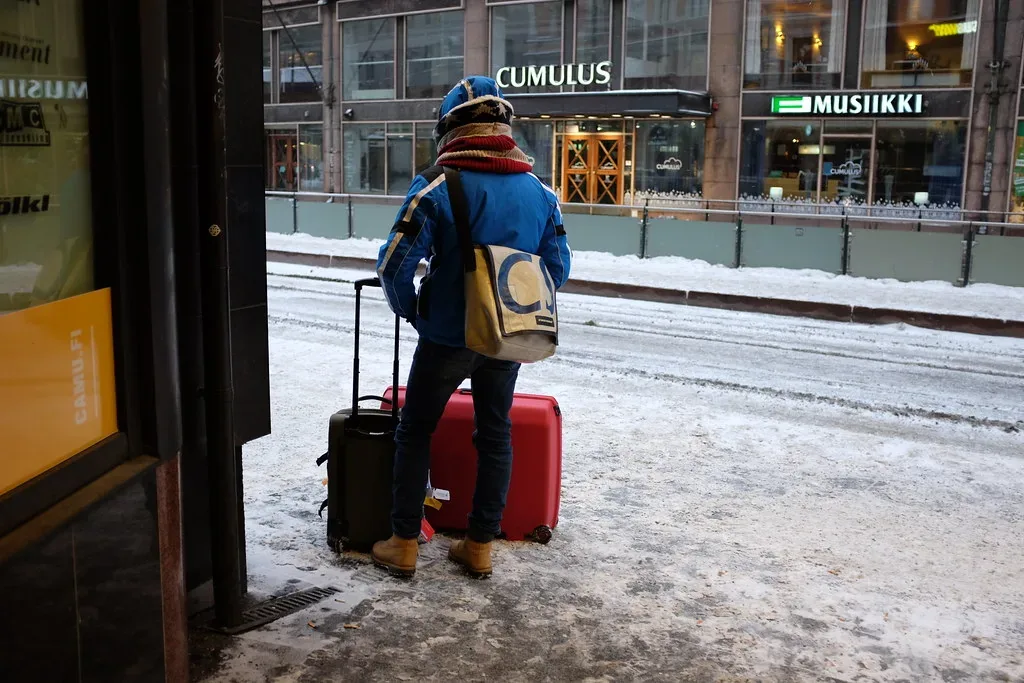 A student standing with a suitcase and backpack at a snowy tram stop after arriving abroad